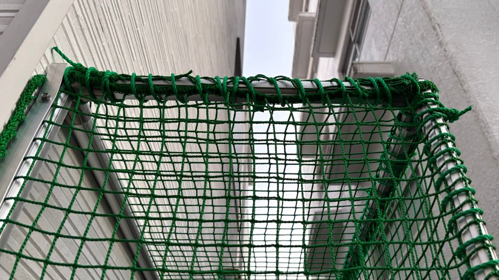 建物外壁の金属フレームに隙間なく頑丈に設置された、緑色のネット施工事例のクローズアップ画像です。