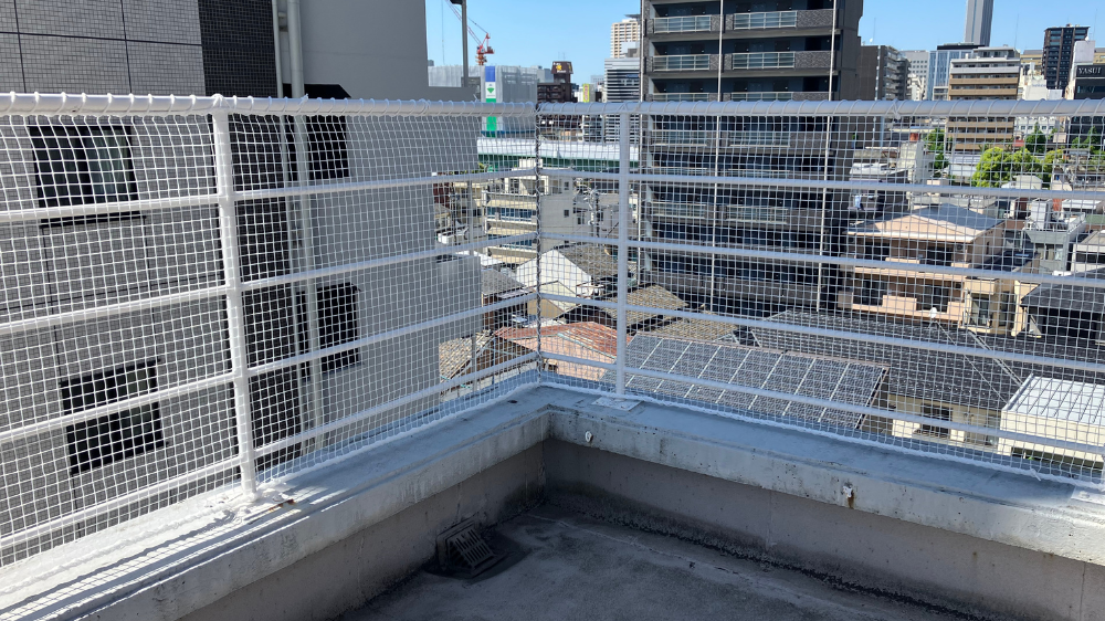 ビルの屋上のコーナーに設置された、市街地を見下ろす白い落下防止ネットとフェンスの全景